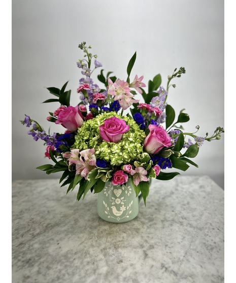 keepsake measuring cup filled with stunning flowers, including pink miniature carnations, Lavender Daiy Pom Poms, Pink Peruvian lilies, and Green Spider Mums
