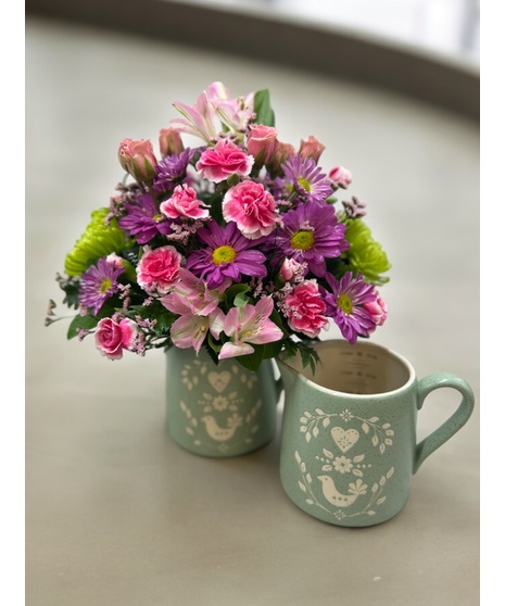 keepsake measuring cup filled with stunning flowers, including pink miniature carnations, Lavender Daiy Pom Poms, Pink Peruvian lilies, and Green Spider Mums