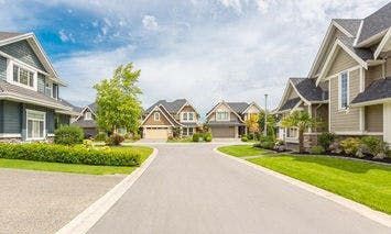 A row of modern suburban houses lines a clean, wide street on a sunny day, with manicured lawns, trees, and gardens adding to a peaceful neighborhood setting.
